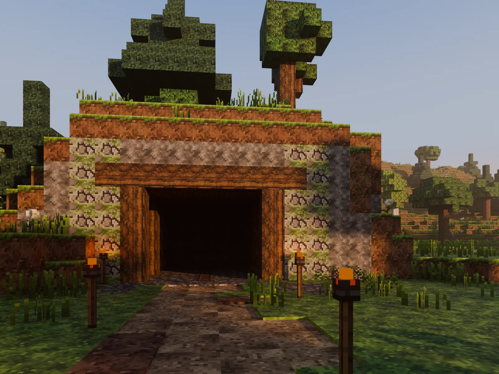 Voxel scene at eye level: a dirt trail lined with torches leads out of the forest to a timber-framed mine entrance in a hillside.
